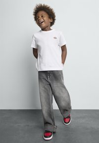 Hvid bomulds T-shirt med logo, grå baggy jeans og røde-sorte sneakers. Barnet står med hænderne bag ryggen foran en lys baggrund.