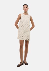 Robe blanche sans manches à texture en dentelle, avec un col rond et un motif floral. Assortie à des ballerines en cuir tressé marron, pour un look décontracté.