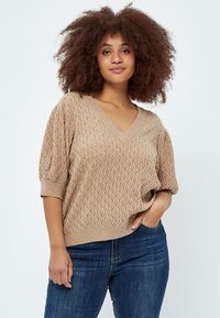 Beige pletený svetr s výstřihem do V, krátkými rukávy a texturovaným diamantovým vzorem. Spárováno s tmavými džíny.