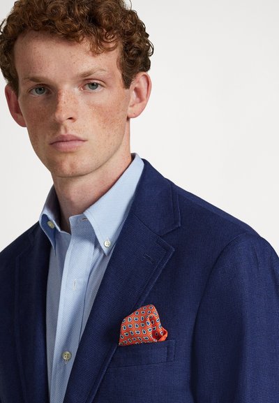 Young man with curly red hair wearing a blue textured blazer, light blue buttoned shirt, and orange patterned pocket square on white background.