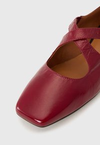 Chaussure plate en cuir rouge avec bout carré et bride croisée sur le cou-de-pied, présentant une finition lisse et une doublure intérieure beige.