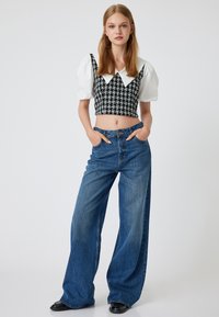 Mustavalkoinen houndstooth-lyhythihainen toppi, jossa on valkoinen kaulus, yhdistettynä vaaleansinisiin leveälahkeisiin farkkuihin. Malli seisoo kädet Taskuissa.