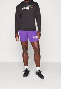 Felpa nera in cotone con un logo colorato, abbinata a pantaloni corti sportivi viola con un logo bianco. Indossata con sneakers nere e calze bianche.