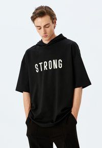 Sudadera con capucha de manga corta negra, confeccionada con tejido suave, que presenta la palabra "FUERTE" en letras blancas y en negrita a lo largo del pecho.