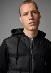 Gilet en doudoune noir avec fermeture éclair, capuche amovible et manches longues. Présente une texture brillante et matelassée avec des détails de fermeture éclair en argent élégant.