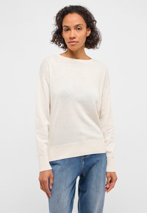 Junge Frau mit lockigem Haar, die einen schlichten weißen Pullover mit langen Ärmeln und blaue Jeans trägt, stehend vor einem einfarbigen weißen Hintergrund.