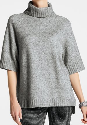 Woman wearing a loose gray knit turtleneck sweater with half sleeves and patterned black and white pants, standing against a white background.