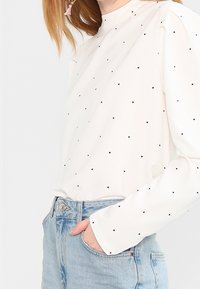 Haut blanc à manches longues avec des pois noirs, doté d'un col montant et d'une coupe décontractée, associé à un jean en denim bleu clair.