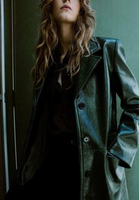 Cappotto in pelle verde scuro con finitura lucida, revers strutturati, grandi bottoni e una vestibilità oversize, indossato sopra una camicia nera.