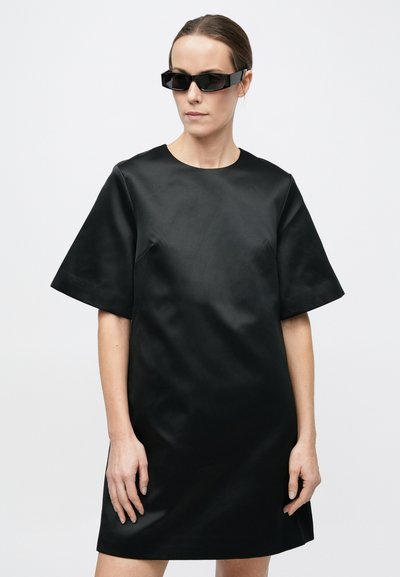 Robe noire à manches courtes en tissu lisse, avec un col rond et une coupe décontractée. Accessoirisée de lunettes de soleil noires à monture angulaire.