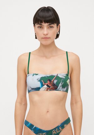 Donna che indossa un costume da bagno a due pezzi con sottili spalline verdi e una grafica di un surfista sulla parte superiore, su uno sfondo bianco semplice.