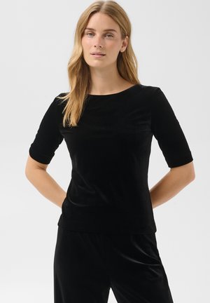 Jeune femme aux longs cheveux blonds portant un haut en velours noir à manches mi-longues et un pantalon assorti, debout, les mains dans le dos.