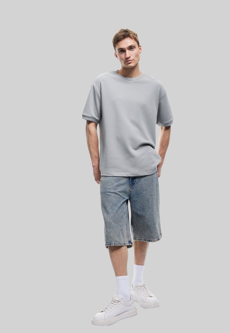 Grå kortærmet t-shirt med en afslappet pasform, sammen med blå denimshorts. Modellen har hvide sneakers og hvide ankelsokker på.