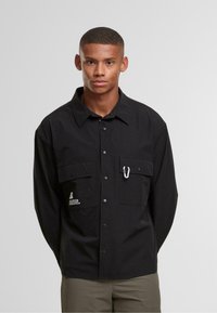 Młody mężczyzna z krótkimi włosami ubrany w czarną koszulę zapinaną na guziki z dwoma kieszeniami na piersi, w jednej z nich znajduje się biały karabinek, stoi na tle o jednolitym kolorze.