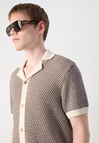Short-sleeved polo shirt in a textured brown and cream pattern with a beige collar and buttons. Model wears dark sunglasses.