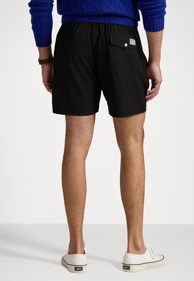 Schwarze Shorts mit elastischem Bund, einer Gesäßtasche mit Druckknopfverschluss und einer glatten Textur, getragen mit weißen Slippern.