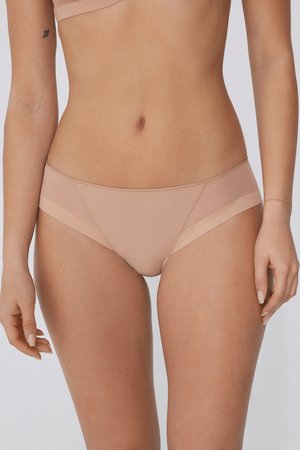 Femme portant des sous-vêtements beiges sans coutures et un soutien-gorge assorti, debout avec les bras détendus le long du corps devant un fond uni.
