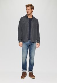 Informele donkerblauwe button-up shirt met een gestructureerd patroon, gecombineerd met lichtblauwe jeans en bruine sneakers. Model staat lachend.