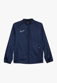 Námořnická modrá větrovka s předním zipem, elastickými manžetami a dvěma bočními kapsami. Na hrudi se nachází bílé logo Nike.