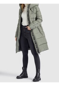 Padded green parka jacket with hood, featuring zippered front, side pockets, and black accents, layered over a white ribbed sweater and black jeans.