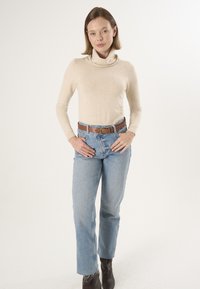 Beige højhalset sweater kombineret med lyseblå lige ben jeans og et brunt bælte; modellen bærer mørkebrune ankelstøvler.