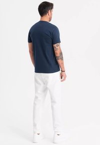 T-shirt blu navy a maniche corte, materiale in cotone, indossato con pantaloni bianchi. Presenta un collo rotondo e tatuaggi visibili sulle braccia.