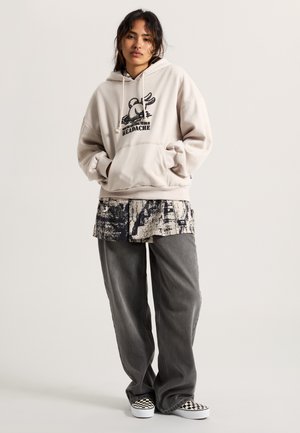 Beige hoodie with graphic print, paired with a patterned, short-sleeve shirt and gray wide-leg pants. Black and white checkered shoes.