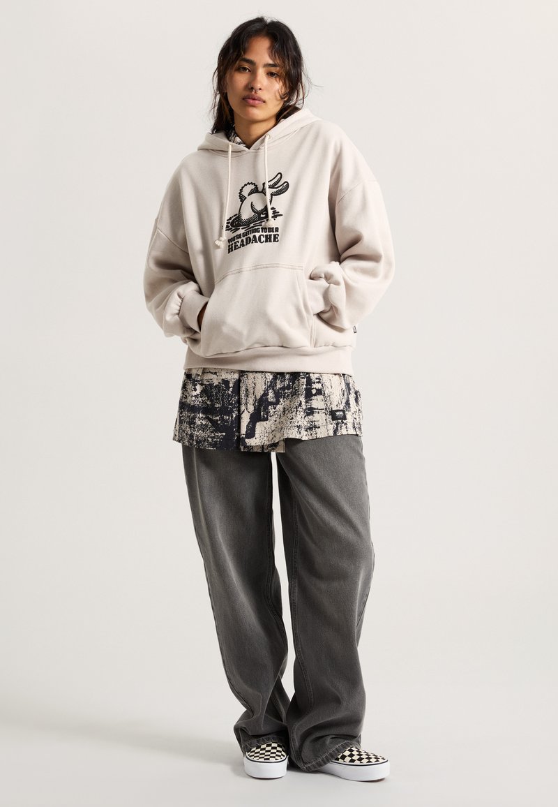Beige hoodie with graphic print, paired with a patterned, short-sleeve shirt and gray wide-leg pants. Black and white checkered shoes.
