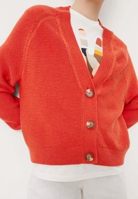 Orange ribbat cardigan med djup V-ringning, tre stora knappar och avslappnad passform, lagd över en vit skjorta med grafisk design.
