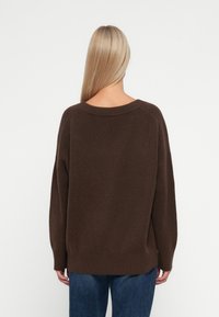 Brun strikket genser med en vid hals, ribbestrikket kant og avslappet passform. Båret over blå denim, som viser en glatt tekstur.