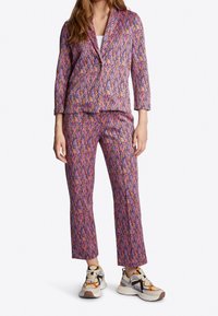 Geprinte blazer en broek in roze, paarse en oranje tinten. De jas heeft een geknipt kraagje; de broek is recht. Gecombineerd met neutrale sneakers.