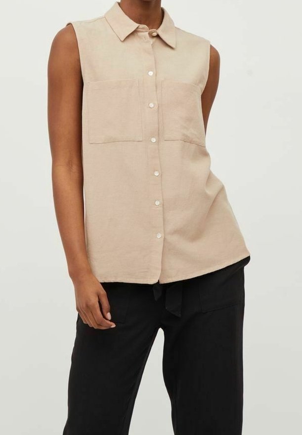 Personne portant une chemise beige sans manches boutonnée avec deux poches poitrine et un pantalon noir ample sur un fond uni.