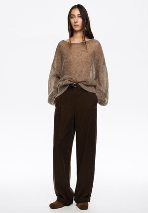 Jasnobrązowy, oversize'owy sweter z dzianiny o półprzezroczystej strukturze, zestawiony z brązowymi, szerokimi spodniami typu wide-leg z paskiem i bocznymi kieszeniami. Brązowe buty.