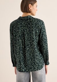 Zielona bluzka w zwierzęcy print o luźnym kroju, z długimi rękawami i detalem w postaci zakładki z tyłu, wykonana z miękkiego materiału. Połączona z szarymi spodniami.