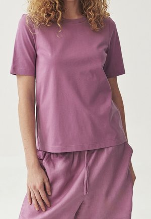 Kobieta w lawendowym t-shircie z krótkim rękawem i pasujących szortach z troczkiem, stojąca z jedną ręką opartą na udzie.