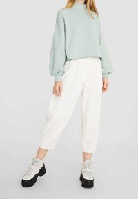 Sudadera verde claro de manga larga y cuello alto; combinada con pantalones blancos holgados y botas gruesas negras y plateadas. Detalles de tela texturizada visibles.