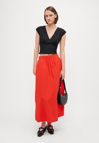 Fekete rövidujjú crop top, élénk piros maxi szoknya és stúdiókkal díszített fekete kézitáska. A modellen fekete blokk-sarkú cipők vannak.