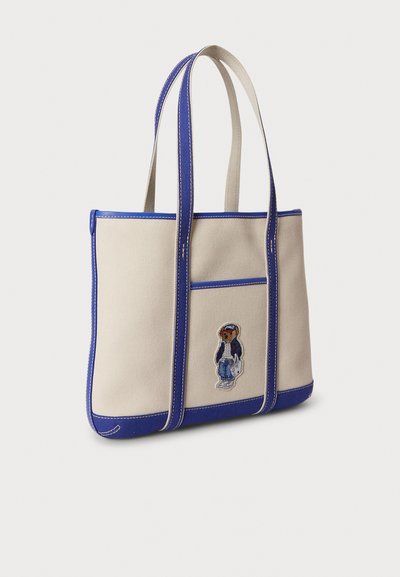 Beige vászon táska kék részletekkel és fogantyúval. Elöl zseb és hímzett karakterdíszítés található. Téglalap alakú és masszív kialakítású.