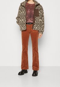 Pantalones de pana marrones de campana, chaqueta de forro polar estampada con cremallera y una camiseta gráfica con un diseño de león. Botas negras completan el atuendo.