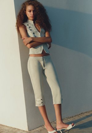 Kvinde med krøllet hår iført lyse denim-korte bukser, ærmeløs knapvest og hvide sandaler med hæl, stående op ad en væg med krydsede arme.