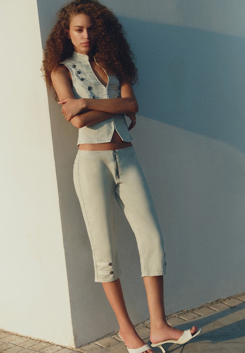 Femme aux cheveux bouclés portant un pantalon court en denim clair, un gilet sans manches boutonné et des sandales blanches à talons, debout contre un mur, les bras croisés.