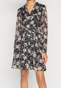 Kvinna i en knälång svart klänning med vita blommor och repmönster, genomskinliga långa ärmar och ett knutet bälte i midjan.