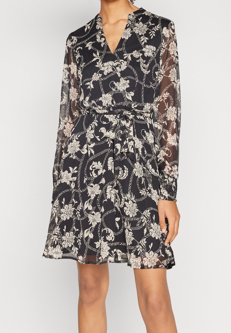 Kvinna i en knälång svart klänning med vita blommor och repmönster, genomskinliga långa ärmar och ett knutet bälte i midjan.