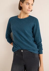 Maglione lungo a maniche di colore verde acqua con un motivo a strisce testurizzato; abbinato a jeans neri con chiusura a bottone e tasche.