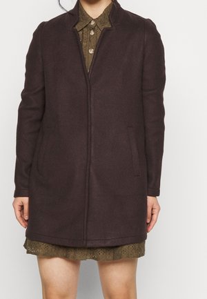 Kobieta ubrana w ciemnobrązowy płaszcz za kolano na oliwkowo-zielonej koronkowej sukience z zapiętym kołnierzykiem, stoi na jednolitym tle.