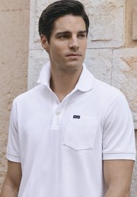 Polo shirt blanc avec un col classique, des manches courtes et une petite poche poitrine avec une étiquette. Textile lisse et design ajusté.
