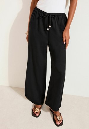 Femme portant un pantalon ample noir avec cordon de serrage et des sandales noires ornées de grandes fleurs beiges, debout devant un fond clair.