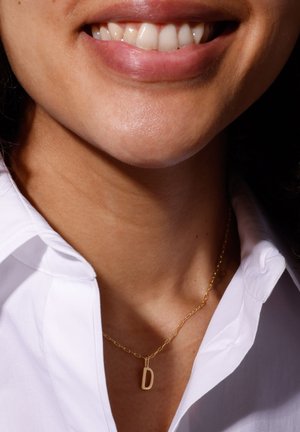 Gouden ketting met een hanger in de vorm van de letter "D", gedragen op een blote nek, tegen een witte overhemd kraag. Glad oppervlak en eenvoudig ontwerp.