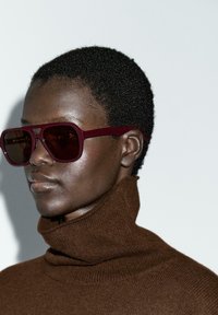 Burgundy oversized sunglasses with dark lenses, square shape, and thick frames worn with a brown high-neck knitted sweater.