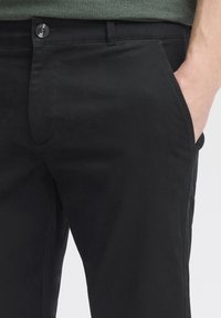 Zwarte katoenen chinos met een knoopsluiting, zijzakken en een soepele textuur. Zichtbare riemlussen en subtiele stikseldetails.
